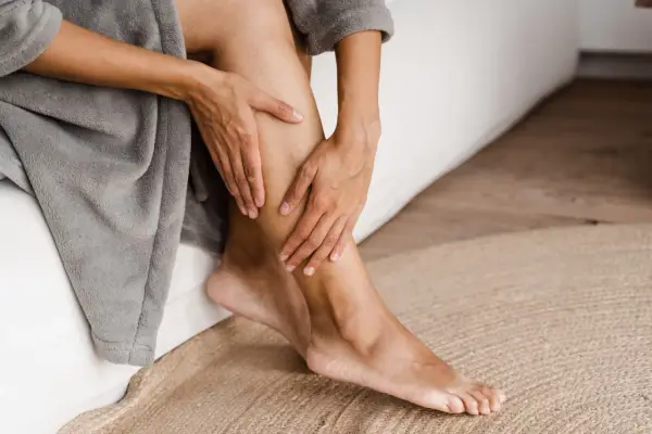 Mani che accarezzano le gambe, evidenziando la pelle secca. La persona è seduta su un letto, avvolta in un accappatoio grigio.