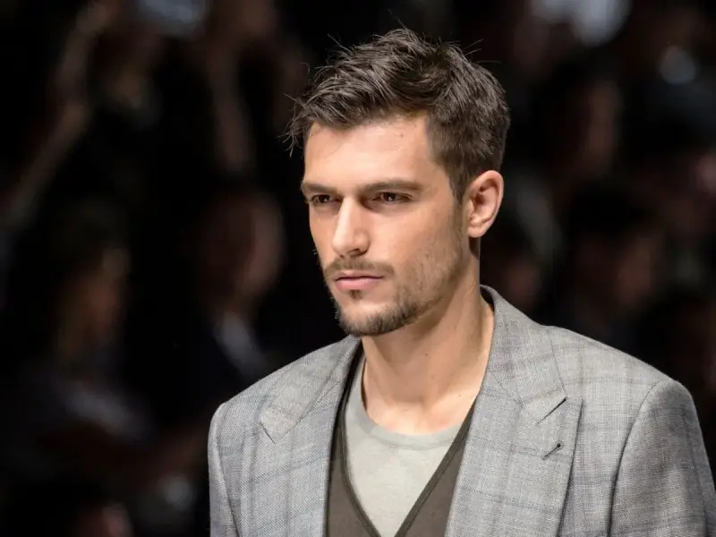 Uomo con capelli spettinati, sguardo intenso, indossa una giacca a quadri grigia.